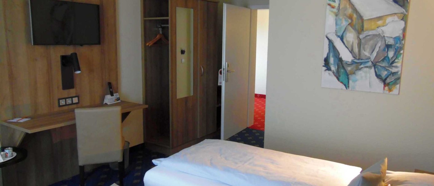 Ein Hotelzimmer mit einem Bett, Schreibtisch, Fernseher und einem abstrakten Kunstwerk an der Wand. Der Raum wirkt modern und aufgeräumt., © TOMAS