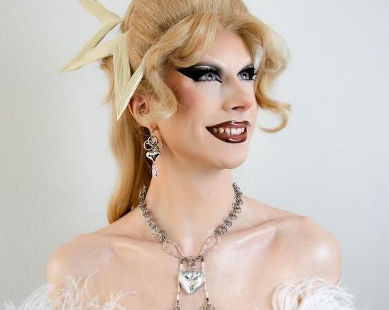 Eine Person mit extravagantem Make-up und Frisur, trägt ein Sternen-Haaraccessoire, weißen Federumhang und auffälligen Silberschmuck, lächelnd zur Seite blickend., © Theaterhaus Stuttgart e.V. Eine Person mit extravagantem Make-up und Frisur, trägt ein Sternen-Haaraccessoire, weißen Federumhang und auffälligen Silberschmuck, lächelnd zur Seite blickend., © Theaterhaus Stuttgart e.V.