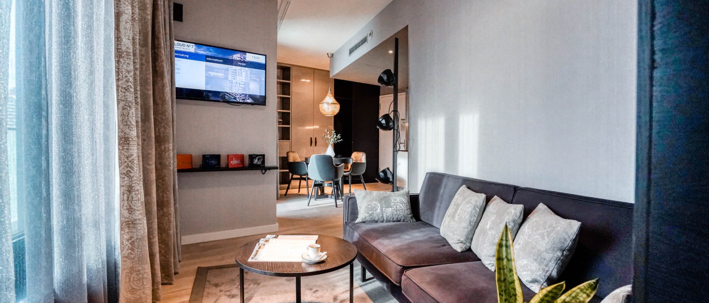Moderne Suite mit elegantem Sofa, Couchtisch und Fernseher. Im Hintergrund ein Essbereich mit Tisch und Stühlen. Dezente Beleuchtung und Vorhänge., © CLOUD N°7 APARTMENTS GmbH