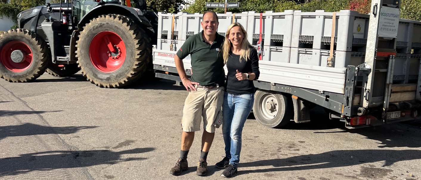 Zwei Personen posieren vor einem Traktor mit Anh&auml;nger voller Kisten. Sie l&auml;cheln in die Kamera. Im Hintergrund sind B&auml;ume und ein Stra&szlig;enschild zu sehen., &copy; weinvomhaigst
