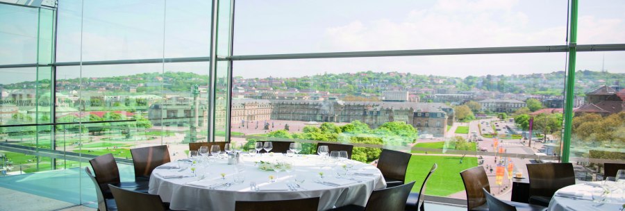 A round table with a white tablecloth and glasses stands in a glassed-in room with a view of a city and historic buildings., © Stuttgart-Marketing GmbH