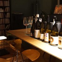 Several bottles of wine stand on a wooden table in a stylish bar. A record shelf and comfortable seating can be seen in the background., © Weinhandlung Kreis KG