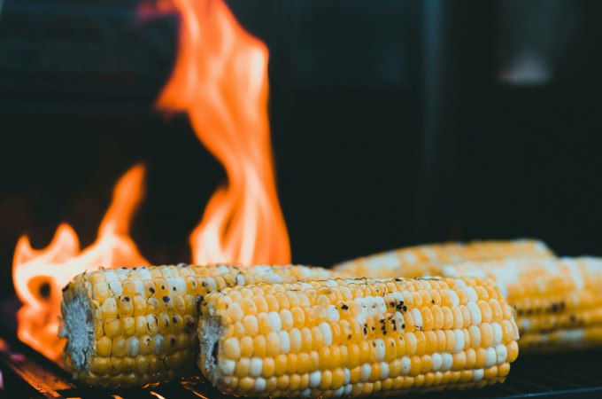 Drei Maiskolben liegen auf einem Grillrost, während im Hintergrund Flammen lodern. Perfekt für ein Veggie BBQ., © Cool-Tours StattReisen Drei Maiskolben liegen auf einem Grillrost, während im Hintergrund Flammen lodern. Perfekt für ein Veggie BBQ., © Cool-Tours StattReisen