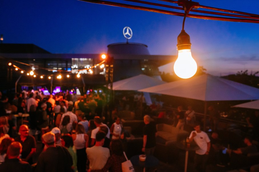 Evening open-air event with lots of people, fairy lights and an illuminated building in the background. A light bulb in the foreground., © Mercedes-Benz Heritage GmbH Evening open-air event with lots of people, fairy lights and an illuminated building in the background. A light bulb in the foreground., © Mercedes-Benz Heritage GmbH