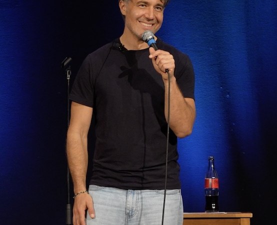 A man on a stage holds a microphone and smiles. Behind him is a blue curtain and a table with a bottle., &copy; Theaterhaus Stuttgart e.V.