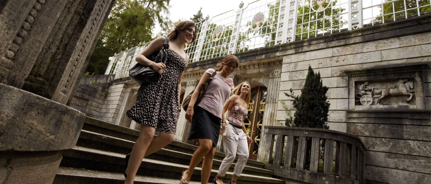 Drei Frauen gehen eine Treppe im Weißenburgpark Stuttgart hinunter, vorbei an einem historischen Gebäude mit Relief und Gitterzaun., © Stuttgart-Marketing GmbH Christoph Düpper