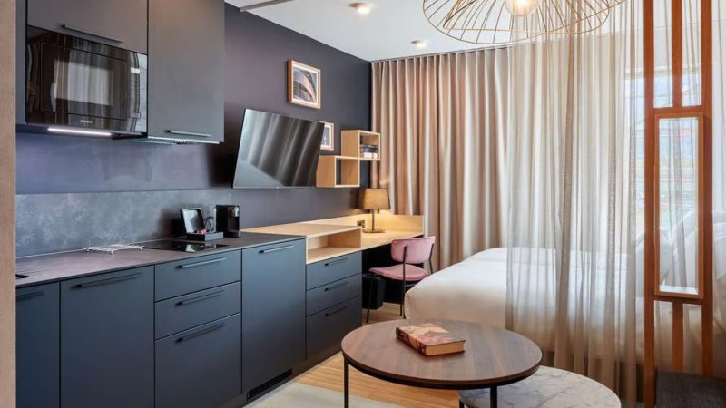 Modernes Studio mit dunkler Küche, Schreibtisch, Bett und stilvoller Beleuchtung. Vorhänge trennen den Schlafbereich ab., © Adagio