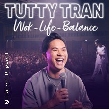 A comedian stands on stage with a microphone, smiling, in front of a large audience. The text "TUTTY TRAN - Wok-Life-Balance" can be seen above it., &copy; links im Bild