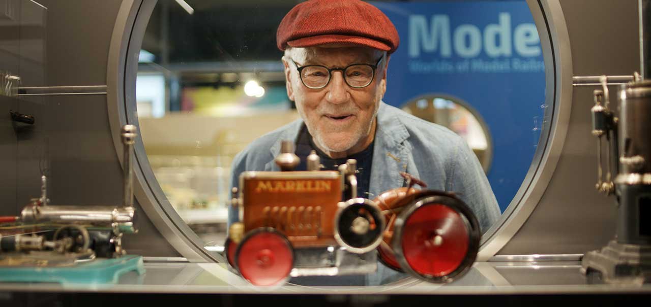 An elderly man in a red hat looks through a round window at a M&auml;rklin model of a tractor in an exhibition., &copy; M&auml;rklineum