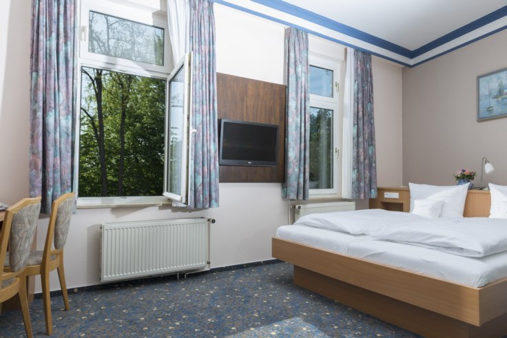 Helles Doppelzimmer mit Bett, Fernseher, Schreibtisch und zwei Fenstern mit Blick auf Bäume. Blau gemusterter Teppich und helle Vorhänge., © Hotel Adler Waiblingen Helles Doppelzimmer mit Bett, Fernseher, Schreibtisch und zwei Fenstern mit Blick auf Bäume. Blau gemusterter Teppich und helle Vorhänge., © Hotel Adler Waiblingen