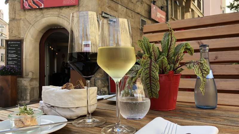 A table with a glass of red and white wine, bread, a small dish and a plant in front of the 'Speisekammer West' restaurant., © Speisekammer West