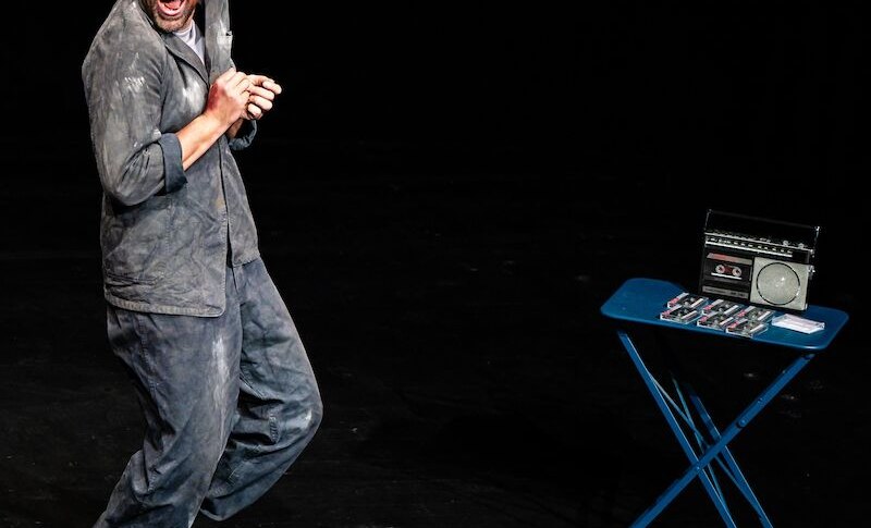 Ein Mann in staubiger Kleidung steht auf einer B&uuml;hne und wirkt begeistert. Neben ihm steht ein Tisch mit einem Radio und mehreren Kassetten., &copy; Theaterhaus Stuttgart e.V.