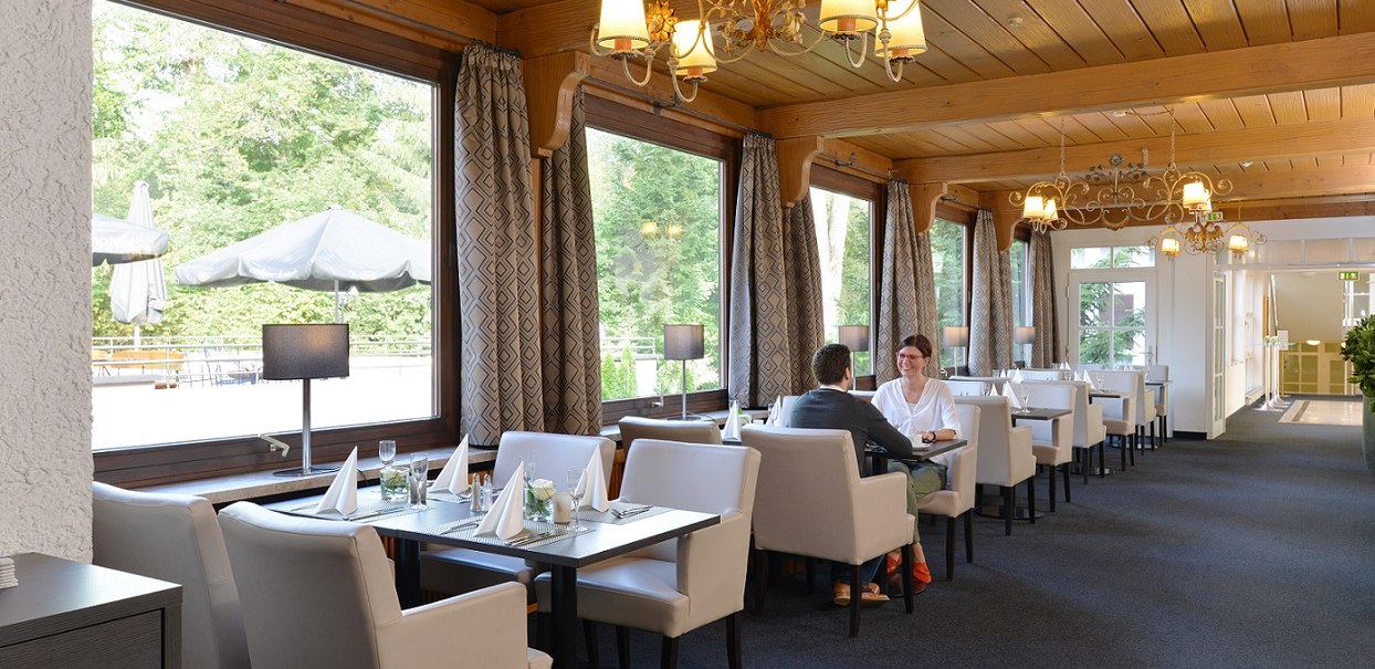 Elegant restaurant with large windows, white armchairs and wooden elements. Two people sit at a table. Sunshades are visible outside., &copy; relexa Waldhotel Schatten