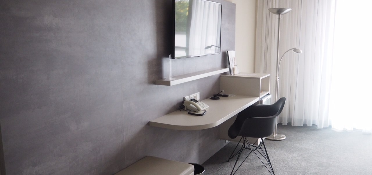 Modern hotel suite with gray desk, black chair, wall-mounted TV and floor lamp in front of a large window with white curtains., © Hotel Fuchsen Modern hotel suite with gray desk, black chair, wall-mounted TV and floor lamp in front of a large window with white curtains., © Hotel Fuchsen