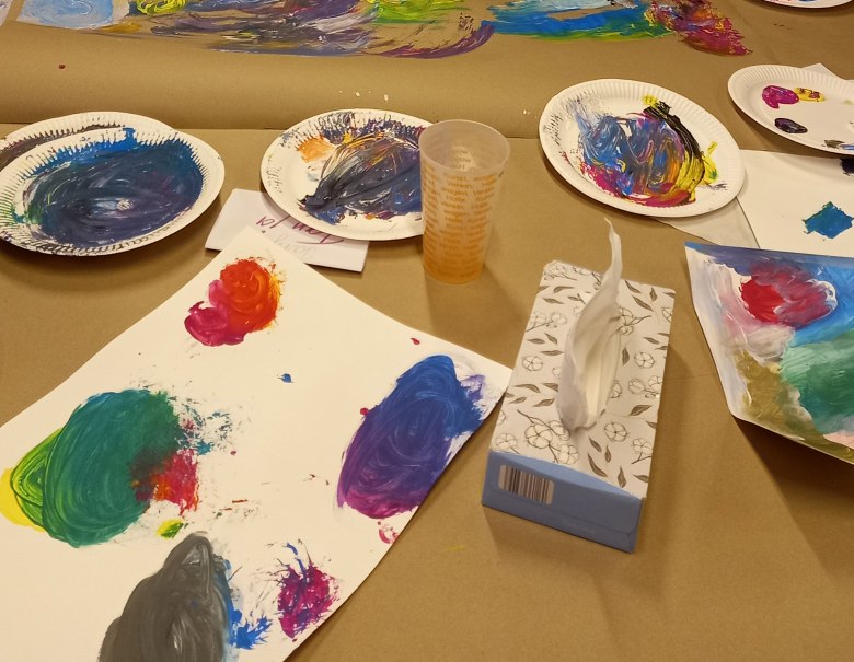 Bunte Farbpaletten und bemalte Blätter liegen auf einem Tisch. Ein Plastikbecher und eine Box mit Papiertüchern sind ebenfalls zu sehen., © KVWH Bunte Farbpaletten und bemalte Blätter liegen auf einem Tisch. Ein Plastikbecher und eine Box mit Papiertüchern sind ebenfalls zu sehen., © KVWH