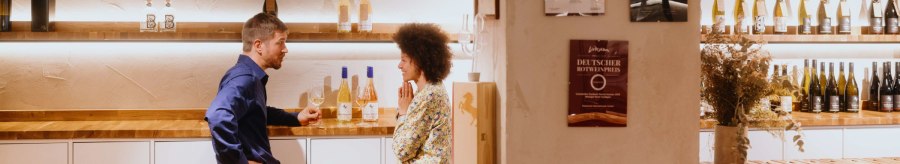 Zwei Personen unterhalten sich in einer Vinothek. Weinflaschen stehen auf Regalen, Urkunden an der Wand. Gem&uuml;tliche Atmosph&auml;re mit Holztischen., &copy; SMG, Alwin Maigler