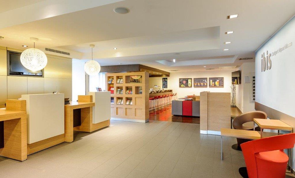 Moderne Hotelrezeption mit Holztheken, Sitzbereich und dekorativen Lampen. Im Hintergrund ein Bücherregal und eine Bar mit roten Hockern., © Ibis Stuttgart Airport Messe