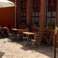 Cozy courtyard with wooden tables and chairs, parasol and plants. Red brick walls and paved floor create an inviting atmosphere., &copy; Gustav Gastro GmbH