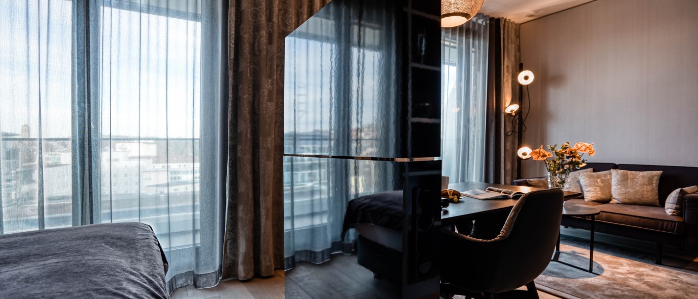 Moderne Suite mit großen Fenstern, eleganten Vorhängen, einem Tisch mit Stuhl, Sofa, dekorativen Lampen und einem Teppich auf Holzboden., © CLOUD N°7 APARTMENTS GmbH