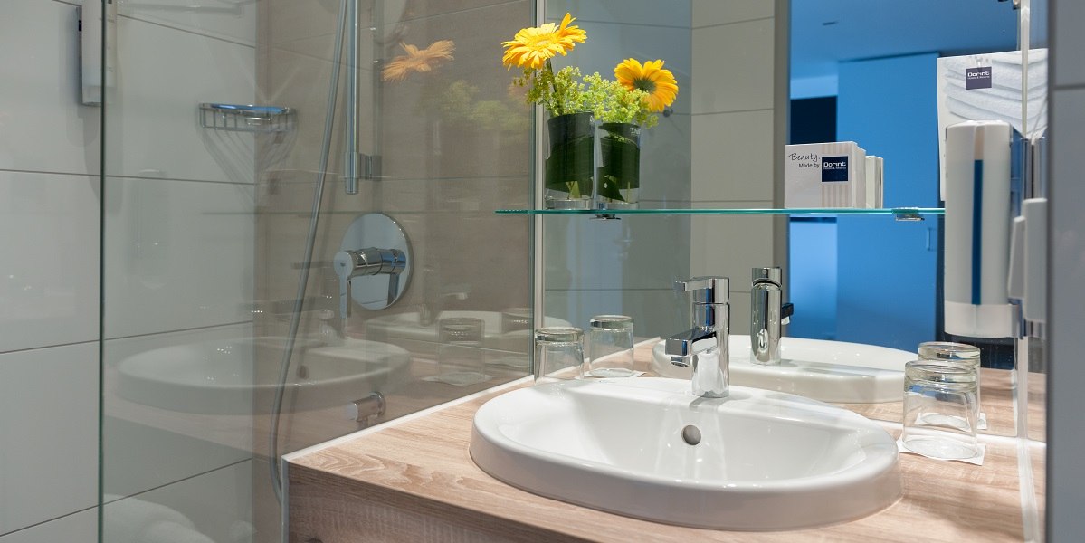 Modernes Badezimmer mit Waschbecken, Spiegel, Glasregal und gelben Blumen. Glasdusche und Pflegeprodukte sind ebenfalls sichtbar., &copy; Essential by Dorint