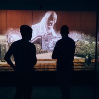 Two people stand in front of a multimedia installation showing Hegel and the Brandenburg Gate. The scene is dark and atmospherically lit., &copy; Julia Ochs