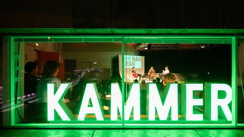 Durch ein Fenster mit der Aufschrift 'KAMMER' sieht man eine Podiumsdiskussion mit mehreren Personen auf einer Bühne., © Württembergische Staatstheater Stuttgart Durch ein Fenster mit der Aufschrift 'KAMMER' sieht man eine Podiumsdiskussion mit mehreren Personen auf einer Bühne., © Württembergische Staatstheater Stuttgart