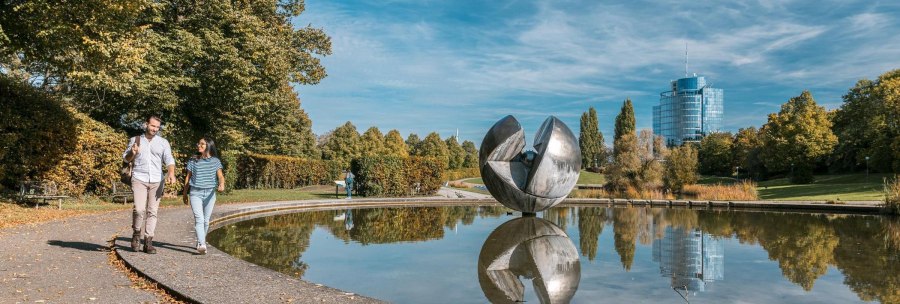 Ein Paar spaziert im Wartbergpark entlang eines Teiches mit einer gro&szlig;en Skulptur und einem modernen Geb&auml;ude im Hintergrund., &copy; Stuttgart-Marketing GmbH, Sarah Schmid