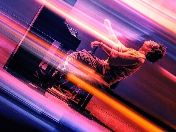 Ein Musiker spielt Klavier auf einer Bühne, umgeben von dynamischen Lichteffekten in lebhaften Farben. Die Szene strahlt Energie und Bewegung aus., © Theaterhaus Stuttgart e.V. Ein Musiker spielt Klavier auf einer Bühne, umgeben von dynamischen Lichteffekten in lebhaften Farben. Die Szene strahlt Energie und Bewegung aus., © Theaterhaus Stuttgart e.V.