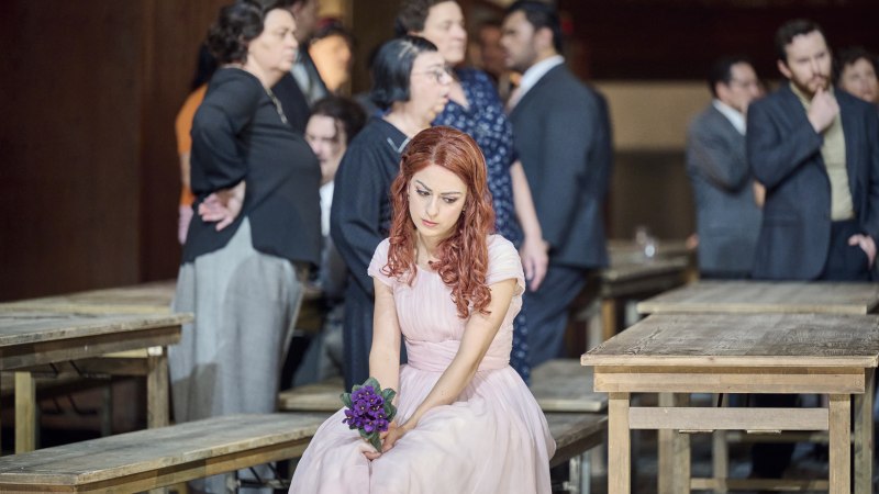 A woman in a pink dress sits thoughtfully on a bench holding a bouquet of flowers. Several people are standing in the background., &copy; Martin Sigmund