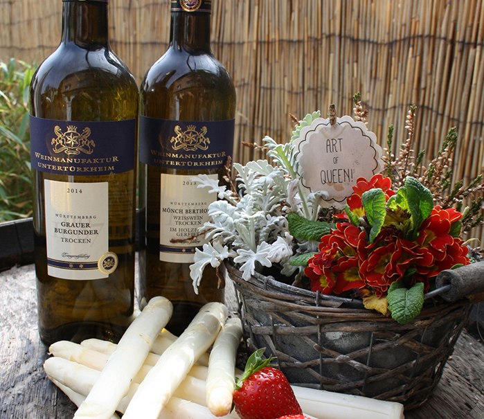 Two bottles of wine, asparagus and strawberries on a wooden table. A flower basket with an 'Art of Queen!' sign stands next to it., © Besenwirtschaft Rauscher