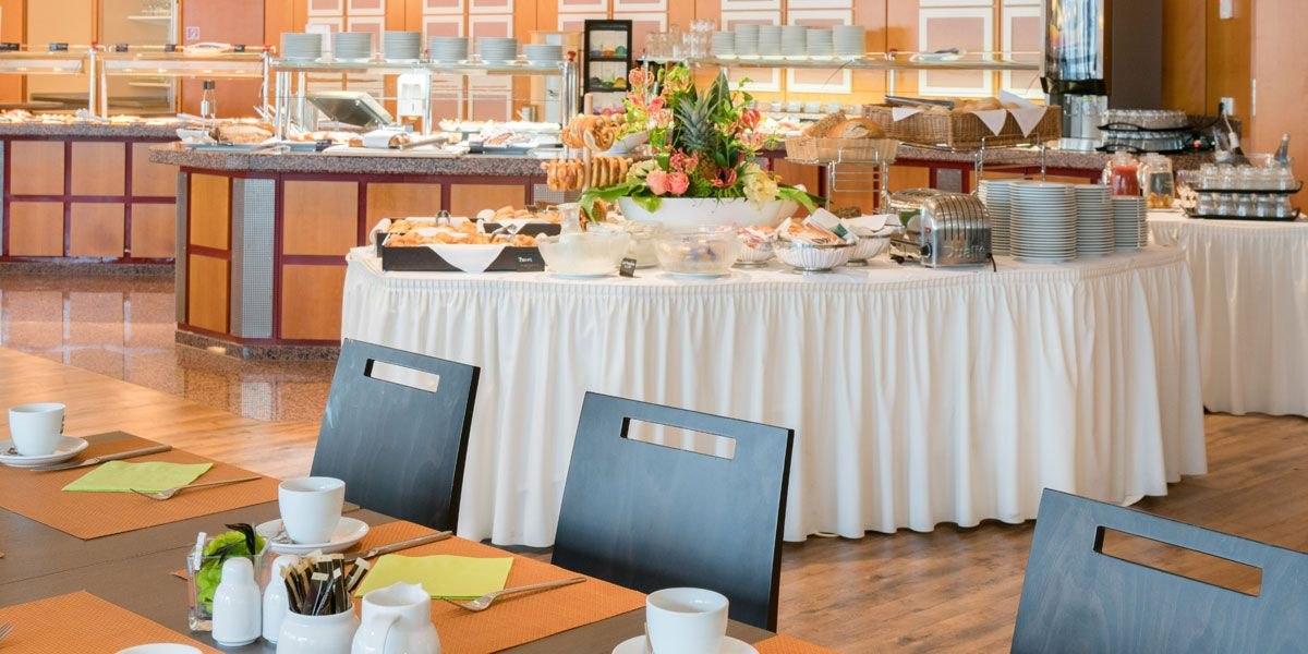 Ein Frühstücksbuffet in einem Hotel mit gedeckten Tischen, Geschirr und einem Blumenarrangement. Verschiedene Speisen sind auf einem Tisch angerichtet., © Best Western Plus Hotel am Schlossberg