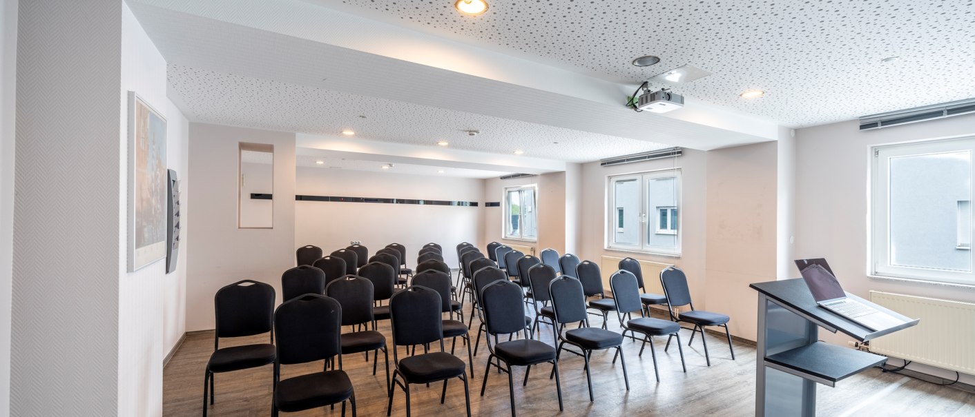 An empty meeting room with several rows of chairs, a lectern and a projector on the ceiling. Bright walls and windows let in light., &copy; Plaza INN Stuttgart-Filderstadt