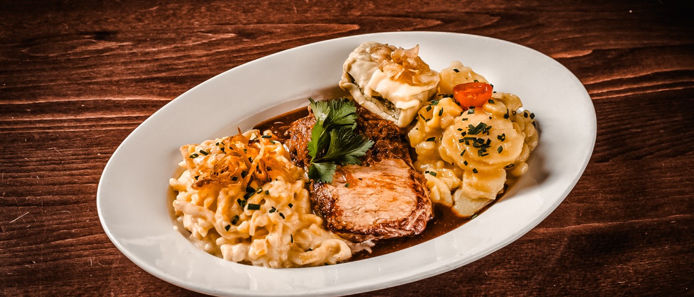 Ein Teller mit Braten, Spätzle, Maultasche und Kartoffelsalat, garniert mit Petersilie, auf einem Holztisch serviert., © Amadeus