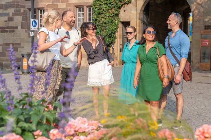 Eine Gruppe von Menschen steht lachend vor einem historischen Gebäude in Stuttgart, umgeben von Blumen und sommerlicher Kleidung., © Stuttgart Marketing GmbH