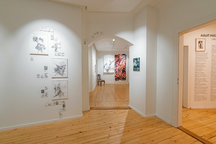 Ein heller Galerieraum mit Holzboden zeigt Kunstwerke an wei&szlig;en W&auml;nden. Ein Stuhl steht im Hintergrund, und ein Text &uuml;ber Adolf H&ouml;lzel ist sichtbar., &copy; Dominique Brewing