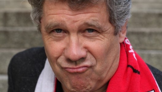 A man with gray hair and a red scarf looks into the camera with a serious expression on his face. Steps can be seen in the background., &copy; Renitenztheater Stuttgart e.V.