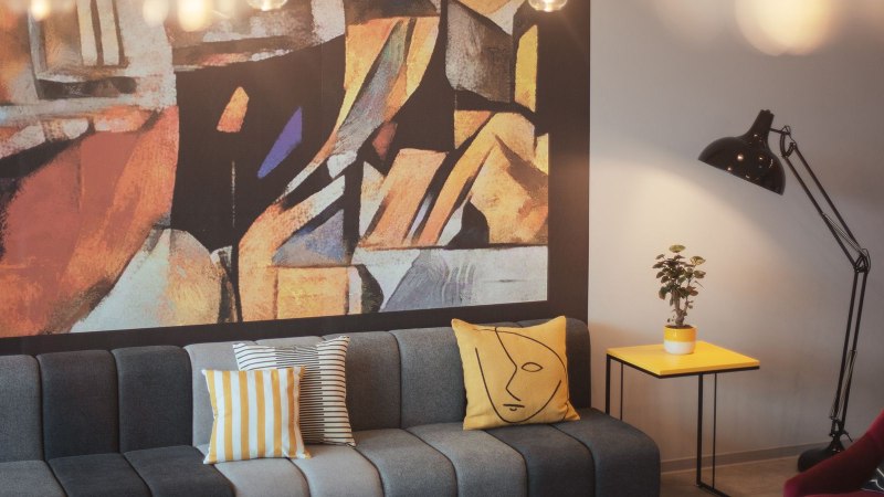 A modern lobby with a gray sofa, colorful pillows, an abstract wall hanging, a yellow side table, a plant, and a floor lamp., &copy; the niu Form - Novum Hospitality
