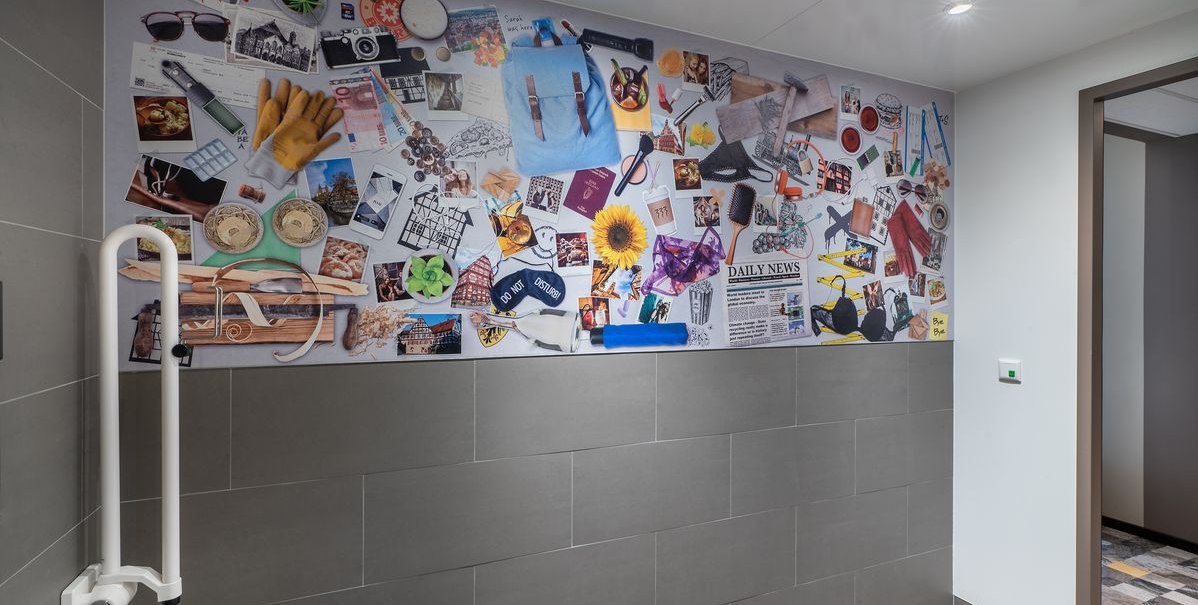 Wand mit bunter Collage aus Fotos und Objekten in einem modernen Raum mit Haltegriff und grauen Fliesen., &copy; Novum Hospitality