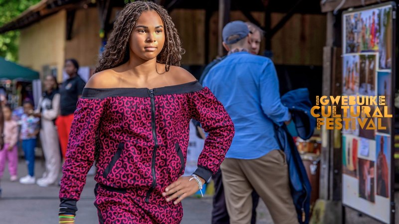 Eine Frau präsentiert ein rotes, gemustertes Outfit auf einem Laufsteg beim Igwe Buike Cultural Festival. Zuschauer im Hintergrund., © igbo Cultural Foundation e.V. Eine Frau präsentiert ein rotes, gemustertes Outfit auf einem Laufsteg beim Igwe Buike Cultural Festival. Zuschauer im Hintergrund., © igbo Cultural Foundation e.V.