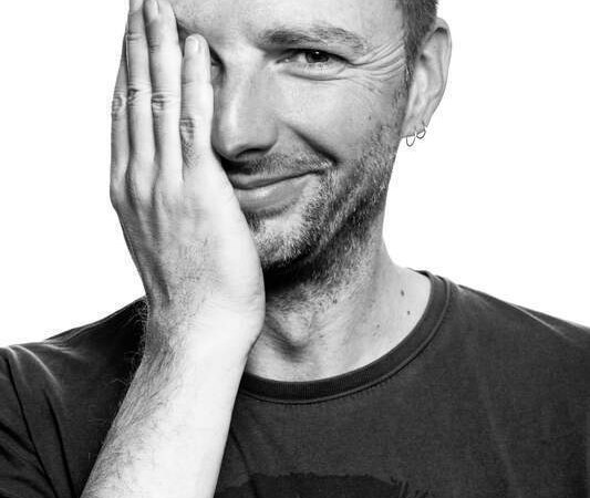 Ein Mann lächelt in die Kamera und hält eine Hand über sein Gesicht. Das Bild ist in Schwarz-Weiß gehalten., © Rosenau Kultur e.V.