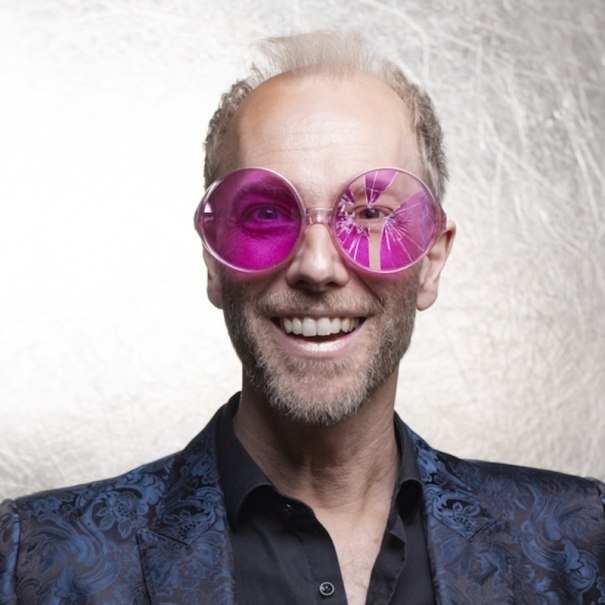 A man wears large pink glasses and a blue patterned jacket against a light background., &copy; Renitenztheater Stuttgart e.V.