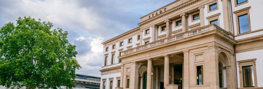 The StadtPalais in Stuttgart has a neoclassical façade with columns. A large tree stands on the left, people sit on the steps., © SMG Thomas Niedermüller
