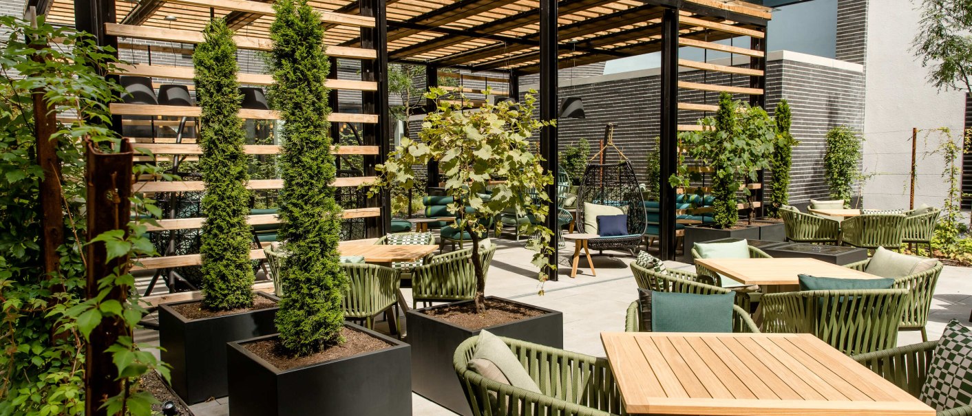 Modern terrace with wooden tables, green chairs and plants. A pergola provides shade, while climbing plants and trees create a green atmosphere., © Motel One - Stuttgart Hauptbahnhof Modern terrace with wooden tables, green chairs and plants. A pergola provides shade, while climbing plants and trees create a green atmosphere., © Motel One - Stuttgart Hauptbahnhof