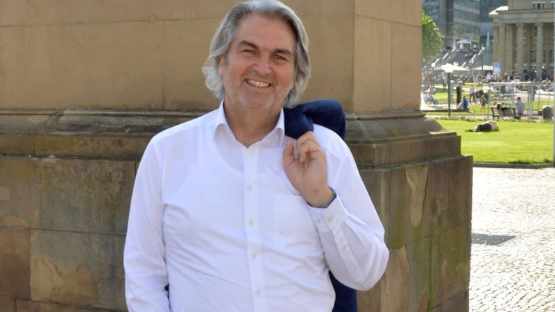 Ein Mann in weißem Hemd steht lächelnd vor einem historischen Gebäude auf einem sonnigen Platz., © Wolfgang Seljé