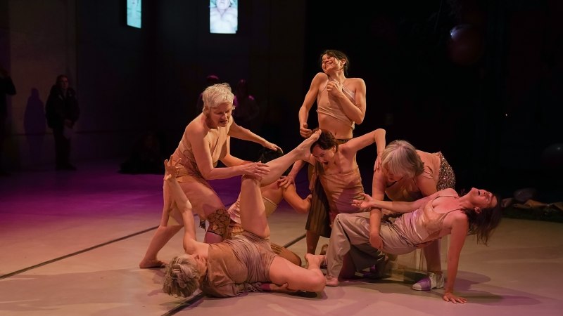 Eine Tanzgruppe in hautfarbenen Kostümen führt eine dynamische Performance auf einer Bühne auf. Die Beleuchtung ist gedämpft und dramatisch., © © Daniela Wolf