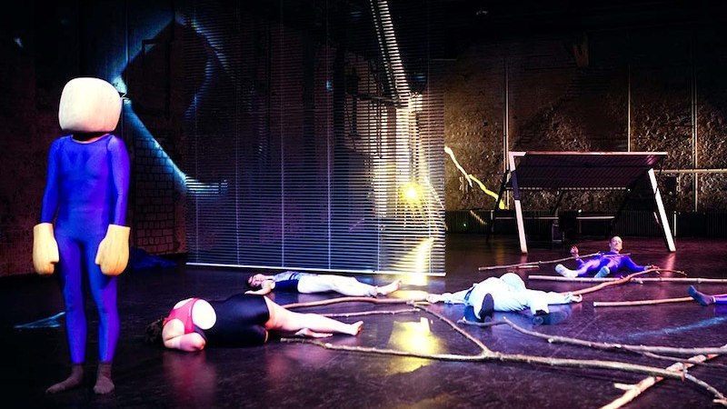 Eine surreale Theaterszene mit einer Figur in einem blauen Anzug und mehreren Personen, die auf dem Boden liegen. Im Hintergrund sind Lichteffekte zu sehen., &copy; Theater Rampe