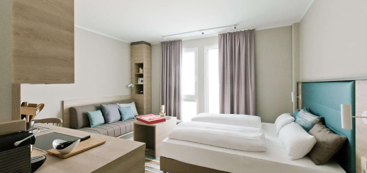Helles Apartment mit Doppelbett, Sofa, Tisch und kleiner K&uuml;chenzeile. Gro&szlig;e Fenster mit Vorh&auml;ngen lassen viel Licht herein., &copy; Arthotel Ana / elaya
