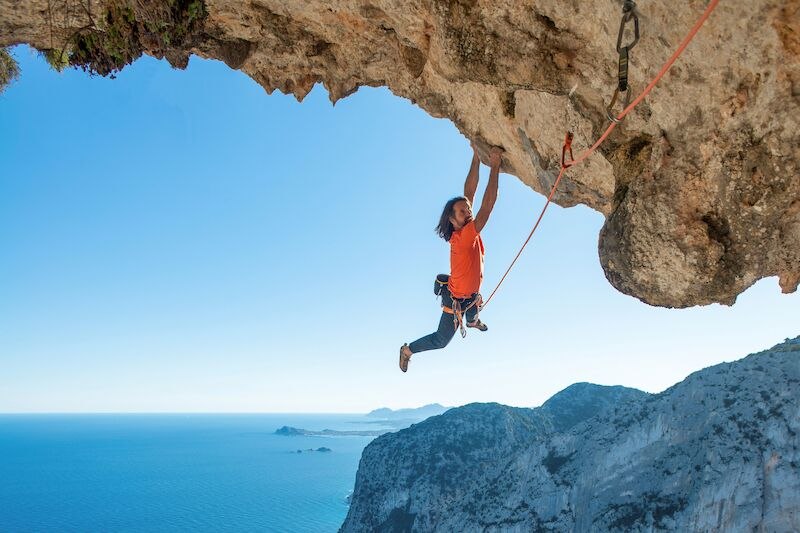 Ein Kletterer in orangefarbenem Shirt hängt an einer steilen Felswand über dem Meer, gesichert mit einem Seil. Im Hintergrund ist das Meer zu sehen., © Theaterhaus Stuttgart e.V. Ein Kletterer in orangefarbenem Shirt hängt an einer steilen Felswand über dem Meer, gesichert mit einem Seil. Im Hintergrund ist das Meer zu sehen., © Theaterhaus Stuttgart e.V.