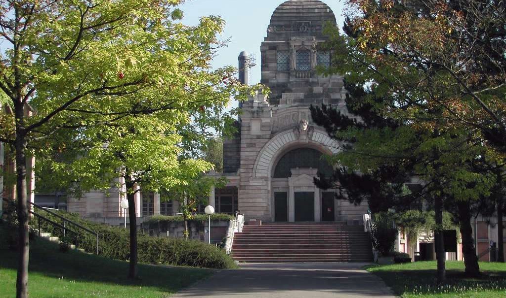Ein historisches Krematoriumsgebäude mit markanter Architektur, umgeben von grünen Bäumen und einem gepflasterten Weg im Vordergrund., © SMG