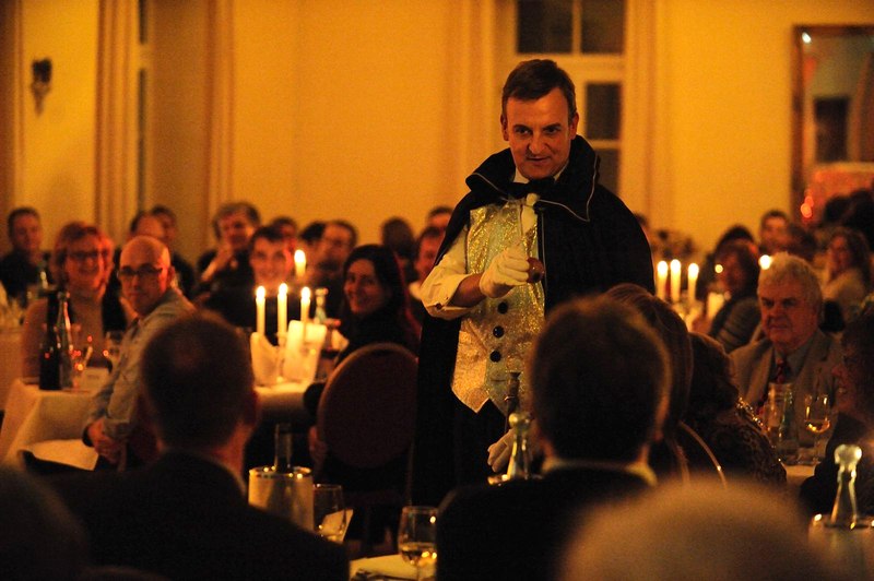 Ein Mann in einem viktorianischen Kostüm spricht zu einem Publikum bei einem Dinner-Event. Der Raum ist in warmes Kerzenlicht getaucht., © Gruseldinner GmbH & Co. KG